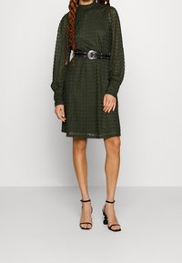 Olivově zelené dlouhé šaty s texturovanou látkou, stažené v pase černým opaskem s kovovou sponou, doplněné černými sandály na tenkých podpatcích.