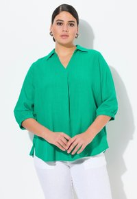 Grøn, let bluse med v-udskæring, optrukne ærmer og plisseret detalje. Stoffet har en subtil tekstur og er parret med hvide bukser.