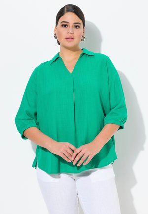 Groene, lichte blouse met een v-hals, opgerolde mouwen en een plooidetail. De stof heeft een subtiele textuur en is gecombineerd met witte broek.