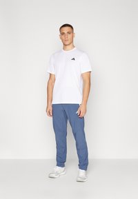 T-shirt blanc à manches courtes avec logo noir et pantalon de sport bleu avec poches latérales, associé à des baskets blanches. Fond neutre.