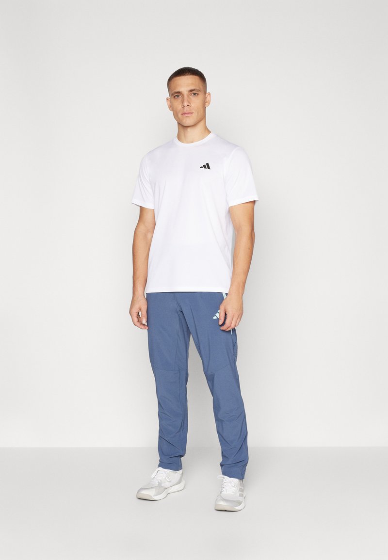 Weißes T-Shirt mit kurzen Ärmeln und schwarzem Logo, kombiniert mit blauen Sporthosen mit seitlichen Taschen und weißen Sneakers. Neutraler Hintergrund.