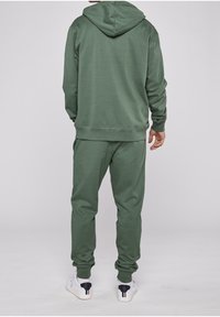 Persona che indossa un felpa con cappuccio verde e pantaloni jogger coordinati, scarpe bianche con dettagli neri sul tallone, in piedi di spalle su uno sfondo bianco.