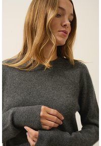 Szary, dziany sweter z prążkowanymi mankietami, okrągłym dekoltem i gładką teksturą. Posiada zapięcie na guzik na ramieniu.