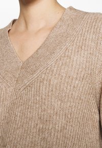 Sweter z dekoltem w serek w jasnobeżowym kolorze, wykonany z dzianiny o fakturowanym wykończeniu. Charakteryzuje się luźnym krojem i ukośnymi przeszyciami przy dekolcie.