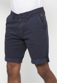 Marineblauwe heren shorts met gerolde zoom, voorzien van voorzakken, ceintuurlussen en een knoopsluiting, gedragen met een wit shirt.