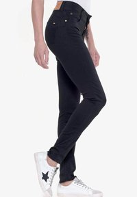 Vaqueros skinny negros con cintura alta, que presentan un tejido texturizado y suave. Los acentos incluyen herrajes dorados y una etiqueta de cuero beige en la parte trasera.