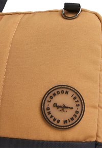Oranžinės tekstūros audinio krepšys su juodu pagrindu, turintis apvalų logotipo pleistrą su užrašu "Pepe Jeans London 1973" ir kilpos prispaudimu.