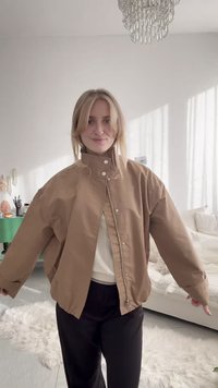 Tânără purtând o jachetă maro supradimensionată, stând într-un living luminos cu pereți albi și un candelabru de cristal.