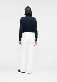 Πουλόβερ σε ναυτικό μπλε με υφή και γιακά, συνδυασμένο με εφαρμοστά λευκά τζιν καμπάνα και μαύρα παπούτσια από γυαλιστερό δέρμα, εμφανιζόμενο από πίσω.