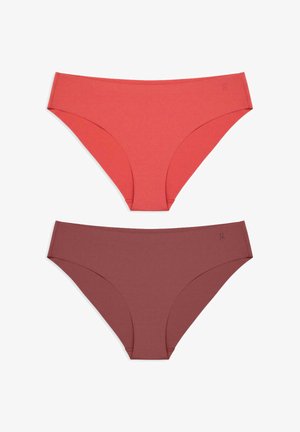 Junge Frau steht barfuß vor einem hellgrauen Hintergrund, trägt ein schwarzes, bauchfreies T-Shirt und braune, hoch geschnittene Unterwäsche.