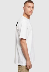 T-shirt blanc en coton avec une coupe ample, un col rond et des manches courtes. Présente un design imprimé sur la manche gauche.