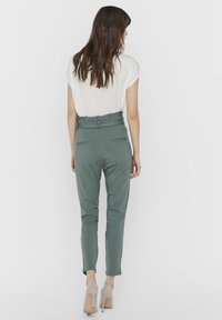 Green tailored pants with a high waistband, slim fit, and two back pockets, paired with a loose, white top. Shoes are light beige.