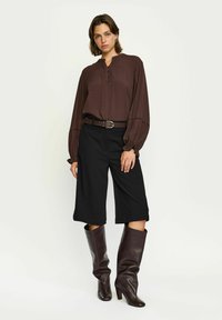 Brun blus med långa ärmar och knappslå, i kombination med svarta vida byxor och knähöga bruna läderstövlar.