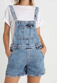 Ljusblå denimöveraller med justerbara axelband, framficka och sidofickor, i kombination med en vit t-shirt.