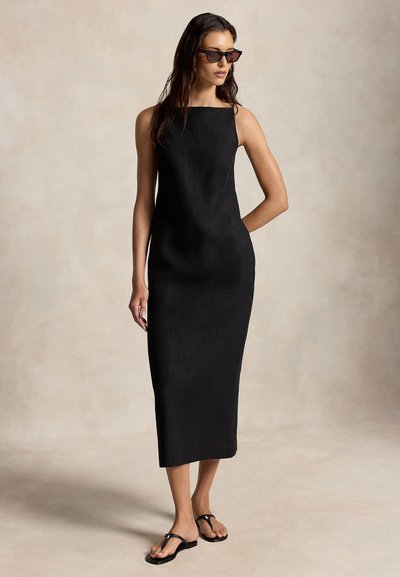 Γυναίκα με μαύρο μίντι αμάνικο φόρεμα, μαύρα γυαλιά ηλίου και μαύρα σανδάλια χαμηλού τακουνιού, που στέκεται μπροστά σε ουδέτερο φόντο.