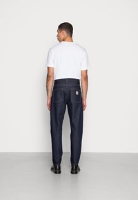Donkerblauwe denim jeans met een rechte snit, voorzien van twee achterzakken en een bruine logo patch. Gekoppeld aan een eenvoudige witte t-shirt.