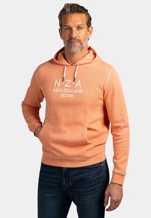 Man met grijs wordend haar en baard, gekleed in een perzikkleurige hoodie met de tekst "N.Z.A. New Zealand 1923" en donkerblauwe spijkerbroek, staand met één hand in de zak.