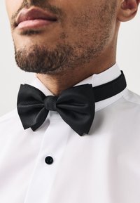 Black textured bow tie with a smooth center knot, worn with a white dress shirt featuring a black button. Close-up view of collar area.