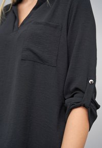 Schwarzes, strukturiertes Hemd mit V-Ausschnitt, einer Brusttasche und hochgekrempelten Ärmeln, die mit Knöpfen befestigt sind, was ein legeres Design betont.