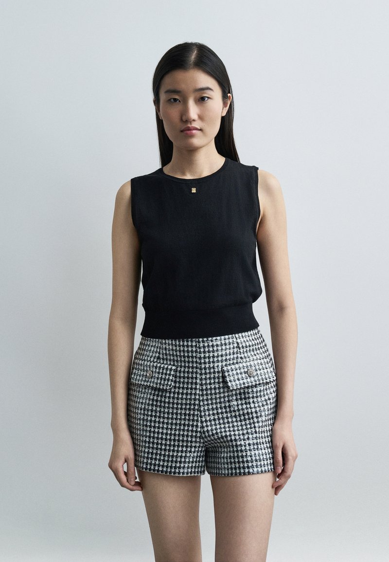 Jeune femme aux longs cheveux foncés portant un haut noir sans manches et un short taille haute à carreaux noir et blanc, face à l'avant sur un fond uni.