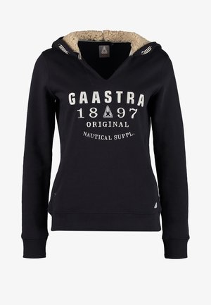 Personne portant un sweat à capuche noir avec un texte blanc, un jean bleu clair déchiré, des baskets bleu marine, et portant un sac à bandoulière en cuir marron.