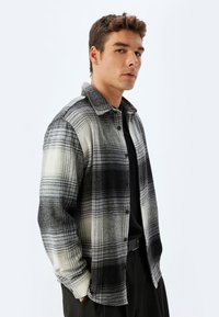 Camisa de franela con un patrón a cuadros en blanco y negro, diseño abotonado y textura suave. Con cuello, de corte relajado y manga larga.