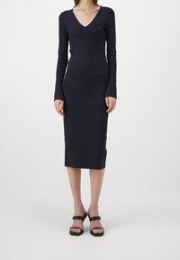 Robe longue bleu marine à manches longues avec un décolleté en V et une silhouette ajustée, présentant un motif diagonal texturé. Une paire de sandales noires avec une double bride.