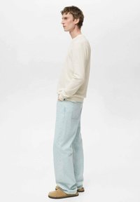 Cămașă bej deschis cu mâneci lungi și textură canelată, asortată cu blugi laterali largi, de culoare albastru deschis, și pantofi slip-on bej, văzuți din lateral.