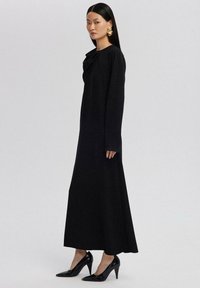 Svart långärmad klänning med draperad halsringning och ett flödande silhuett. Kombinerad med svarta spetsiga klackar. Slätt, enhetligt tyg.