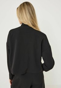 Haut noir à manches longues et coupé, en tissu lisse, avec un col montant et une coupe décontractée, des épaules tombantes et sans motifs visibles.