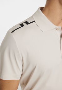 Beige polo-shirt med fast krage, har svarta accentlinjer på axeln, tillverkad av ett mjukt och lätt tyg.
