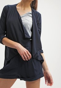 Marinblå omlottjacka i lätt tyg med en avslappnad passform och trekvartslånga ärmar, över en grå t-shirt och shorts.