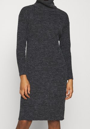 Femme portant une robe-pull en maille côtelée à col roulé, manches longues, de couleur gris foncé, debout devant un fond uni.