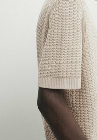 Beige knitted sweater with a textured pattern, short sleeves, and ribbed cuffs. Visible seam on the side, cotton or wool blend.