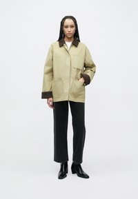 Jachetă utilitară bej cu guler maro, având două buzunare frontale. Asortată cu blugi drepti în culoare închisă și cizme negre până la gleznă.