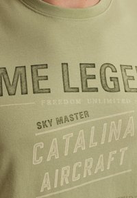 T-shirt en coton vert olive à manches courtes, ornée de graphiques textuels en noir et blanc, incluant "SKY MASTER" et "CATALINA AIRCRAFT."