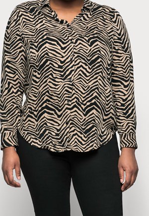 Femme portant une chemise beige et noire à rayures zébrées, boutonnée, manches longues et un pantalon noir, montrée du cou aux hanches.