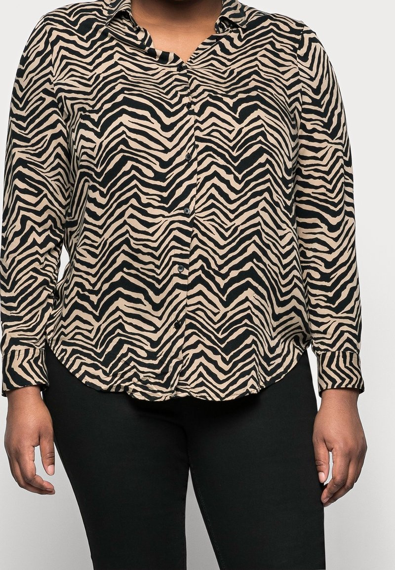 Femme portant une chemise beige et noire à rayures zébrées, boutonnée, manches longues et un pantalon noir, montrée du cou aux hanches.