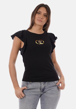 Donna che indossa una maglia nera con maniche arricciate e logo dorato, jeans grigio chiaro, in piedi con le mani nelle tasche su uno sfondo semplice.