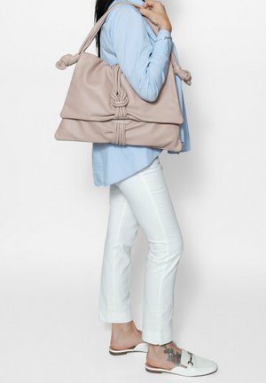 Tan leather handbag with knotted handle, structured shape, and front pleating detail. Model wearing light blue shirt and white pants.