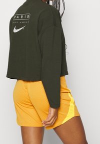 Vihreä lyhythiha sweatshirt, jossa lukee "PARIS" ja Nike-logo. Yhdistetty keltaisiin shortseihin, joissa on tekstuuri ja erottuva sivuvalkoinen raita.