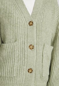 Zelený pletený kardigan s výstřihem do V, třemi velkými hnědými knoflíky, dvěma předními kapsami, texturovanou tkaninou a žebrovanými okraji.
