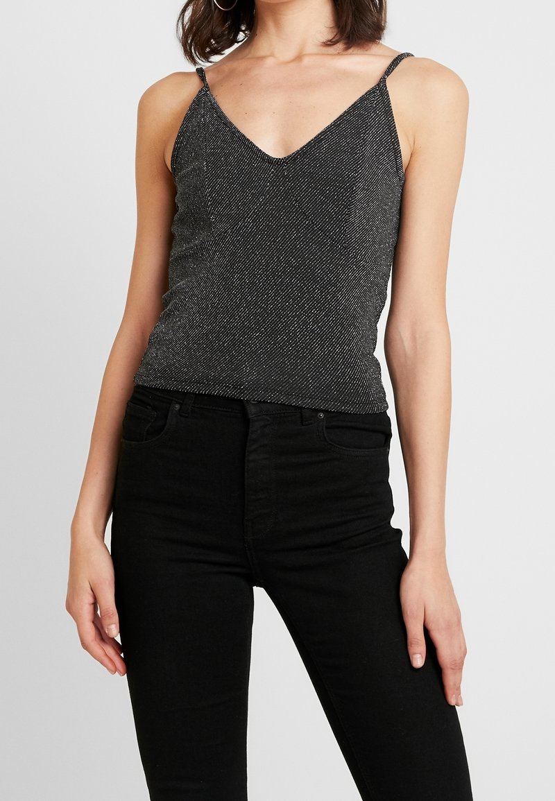 Femme portant un haut noir scintillant sans manches à col en V avec fines bretelles, et un pantalon noir taille haute sur un fond clair uni.