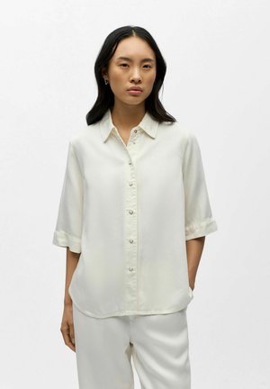 Frau trägt ein locker sitzendes weißes Hemd mit halblangen Ärmeln und passende weiße Hose, steht vor einem einfarbigen weißen Hintergrund.