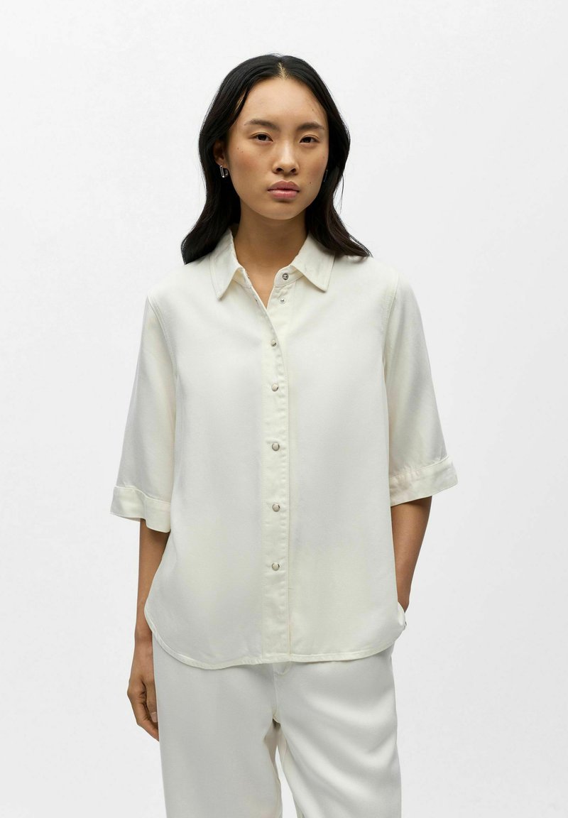 Femme portant une chemise blanche ample à boutons avec des manches jusqu'au coude et un pantalon blanc assorti, debout devant un fond blanc uni.