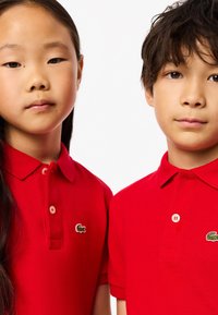 Två barn som bär ljusgröna pikétröjor med en grön krokodil-logotyp, stående nära varandra mot en vit bakgrund.