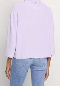 Lila gerippter Pullover mit hohem Kragen und Dreiviertelärmeln, versehen mit einem kleinen Logo-Detail auf der Rückseite. Getragen mit hellblauer Jeans.