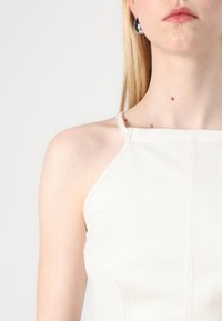 Cream halter top, featuring a structured design with herringbone texture, thin straps, and contrasting stitching. Silver hoop earrings visible.