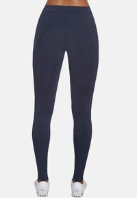 Granatowe legginsy z wysokim pasem, o gładkiej fakturze, z zakrzywionymi przeszyciami i odblaskowymi szwami, w zestawieniu z białymi butami sportowymi.