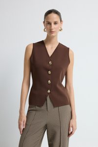 Donna che indossa un gilet senza maniche marrone con bottoni dorati decorativi e pantaloni a vita alta a quadri, in piedi davanti a uno sfondo bianco semplice.